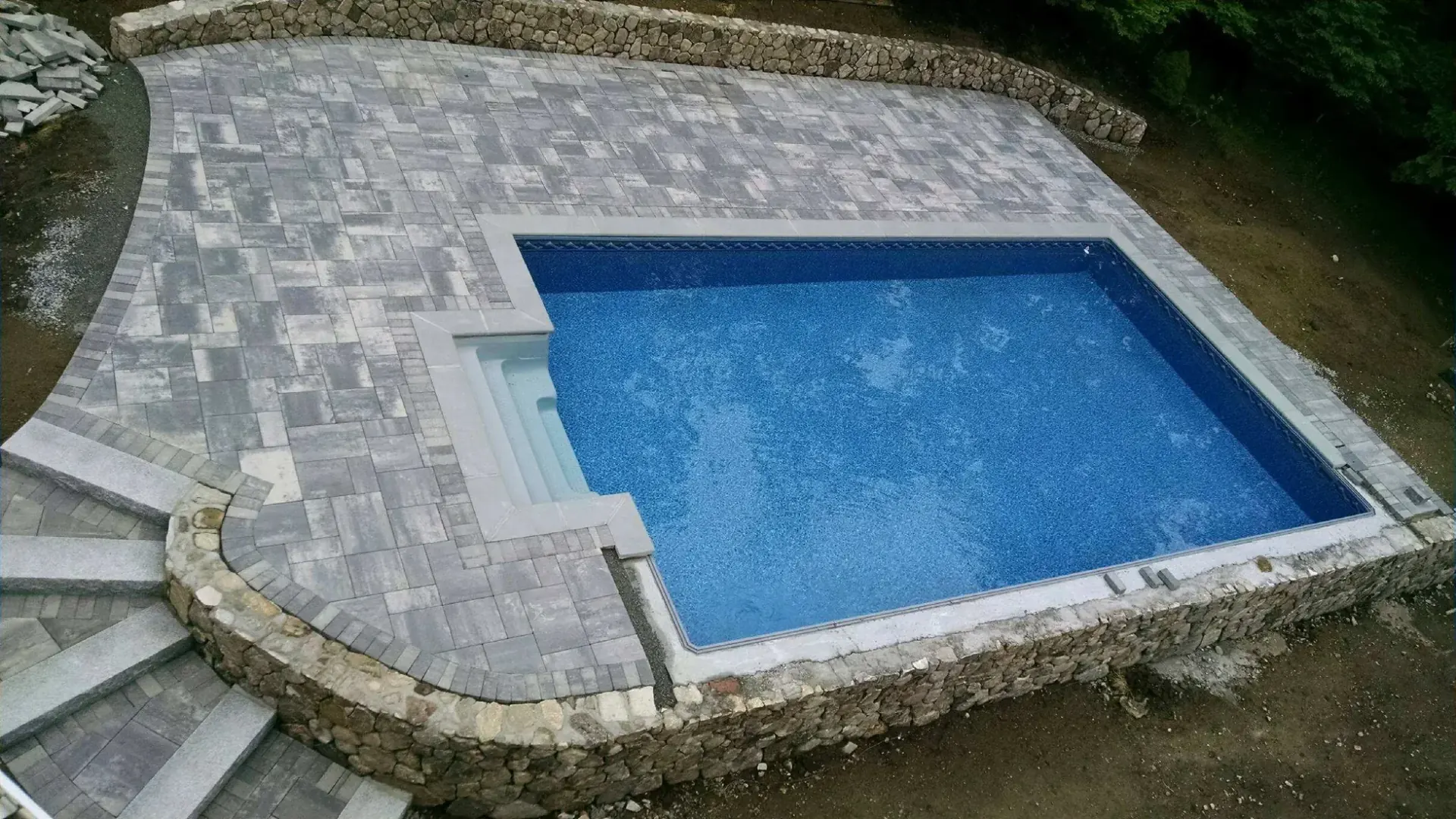 Aerial view of a finished pool deck wrapped in a round-fieldstone retaining wall with granite steps in North Reading, MA