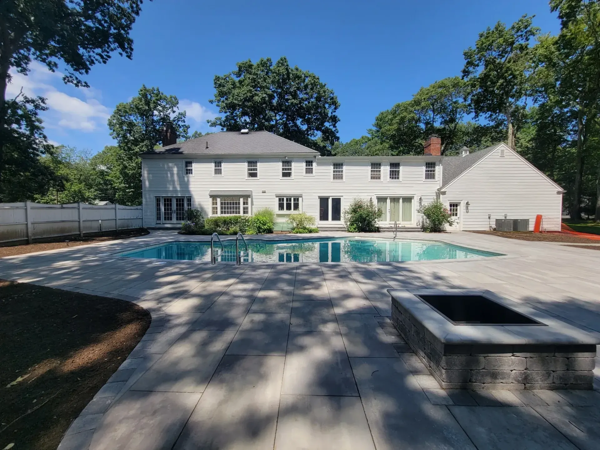 Large-format paver pool deck with a square stone fire pit behind a white colonial home in Lynnfield, MA