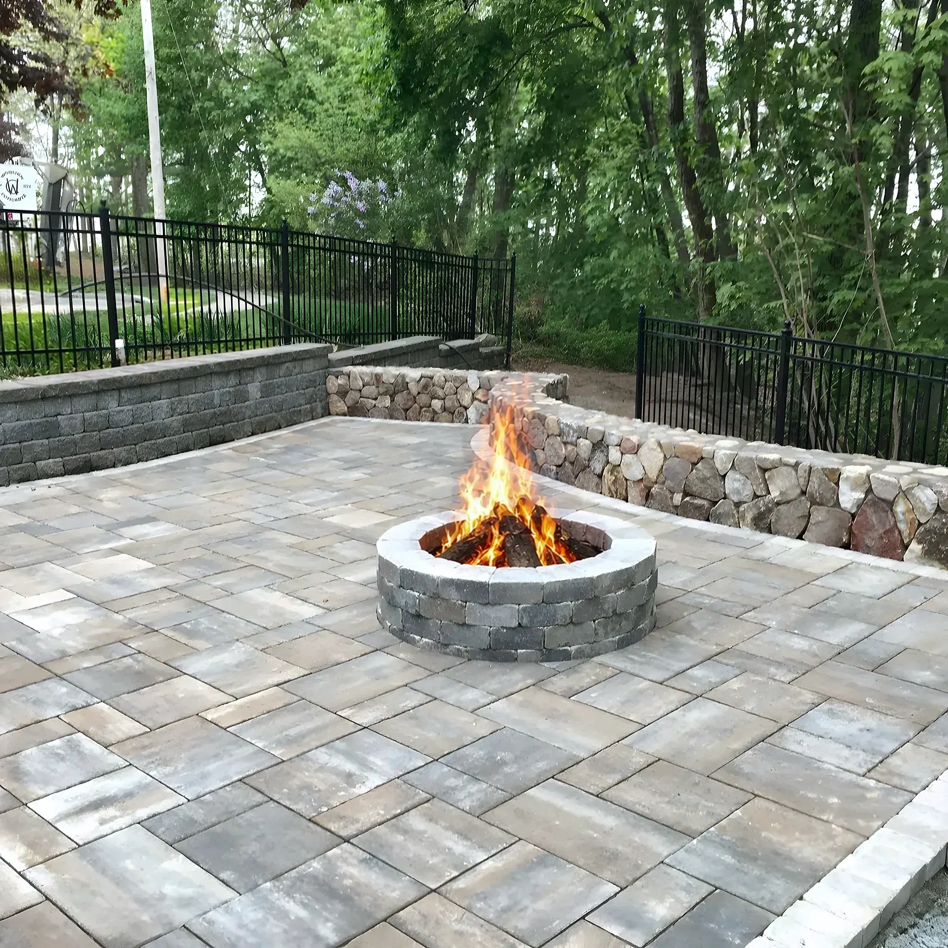 Paver patio with a round stone fire pit and fieldstone seat wall behind a black fence