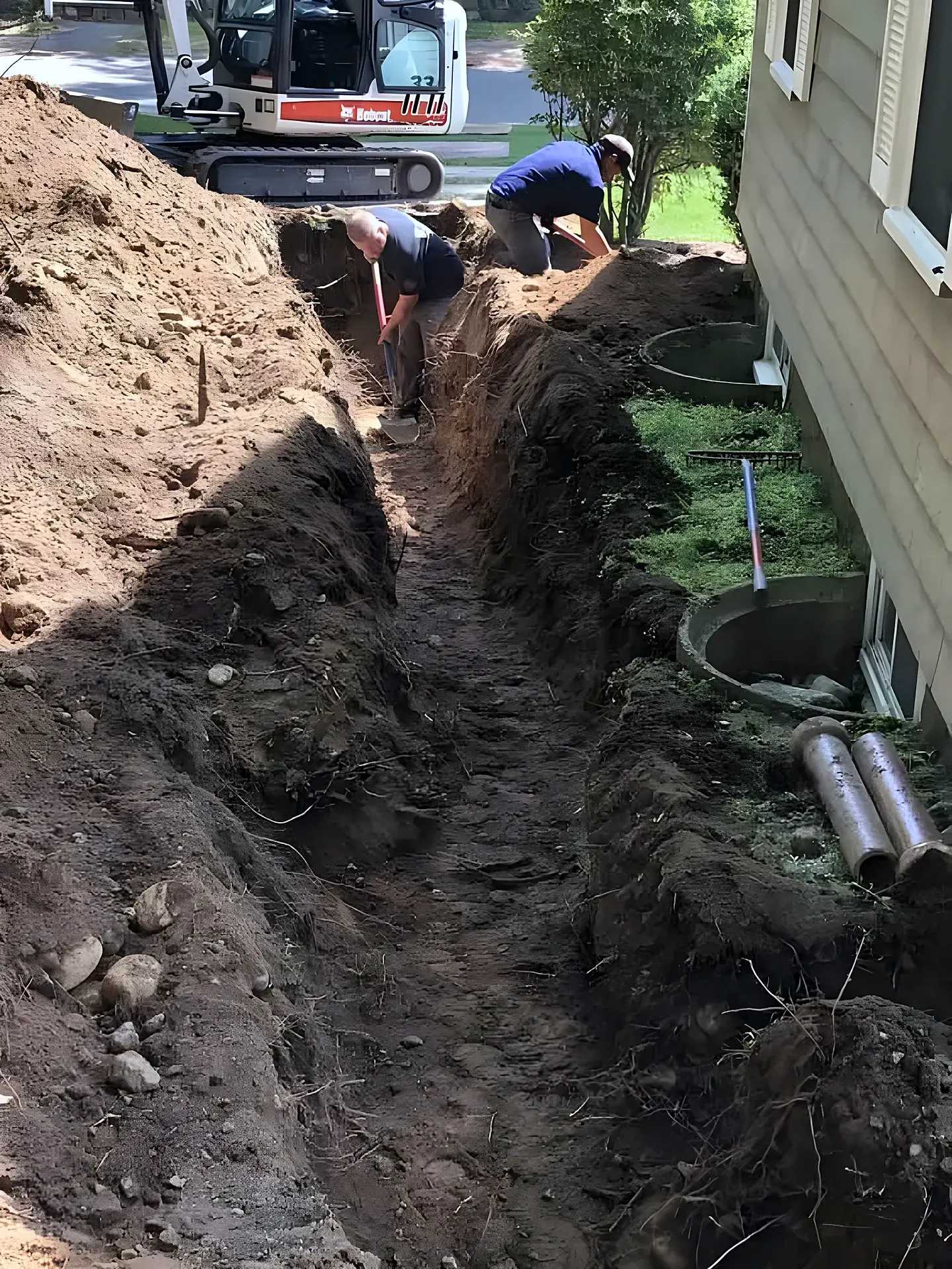 A narrow drainage trench dug along the side of a house, with a small excavator in the background