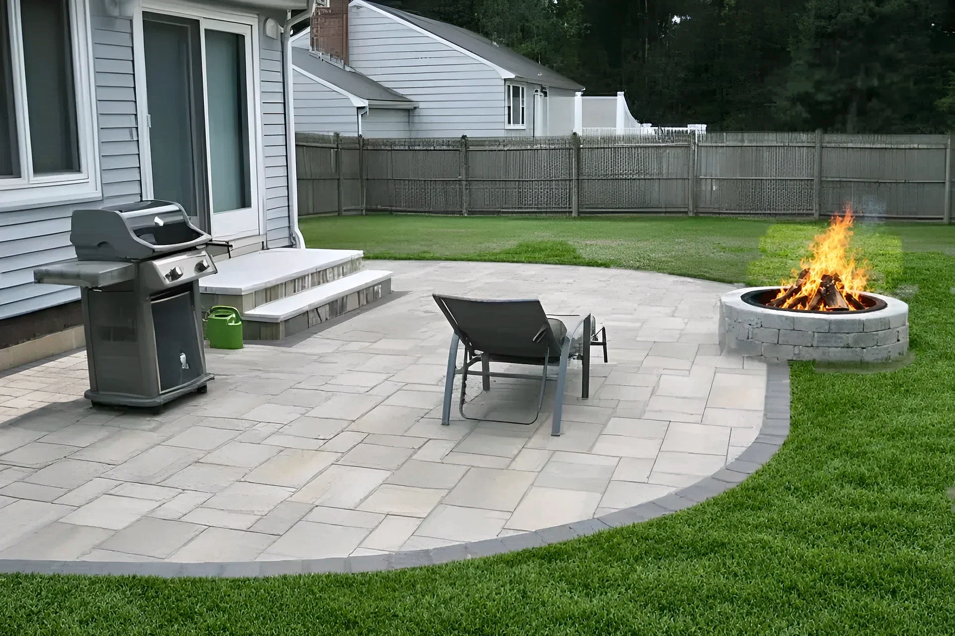 Round block fire pit on the edge of a paver patio with a grill and patio chair